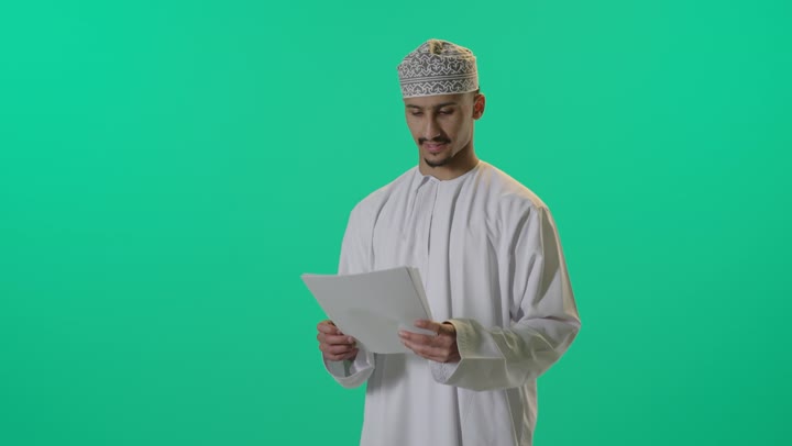 مراجعة نتائج الاجتماع، بورتريه لرجل عربي خليجي عماني يرتدي الدشداشة والكمة يحمل بيده كراسه تدوين الملاحظات يقوم بتصفح الاوراق، اعداد تقارير ومهام العمل، دراسة جدوى الاعمال, خلفية خضراء