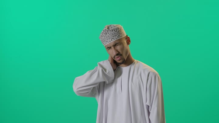 رجل عربي خليجي عماني يرتدي الدشداشة البيضاء و العمامة , تعابير الشعور بالالم و الاعياء, وضع اليد على الرقبة علامة على الوجع الشديد , خلفية خضراء