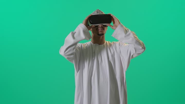 Entertainment and leisure in free time, using modern advanced technologies to watch movies and simulate the imaginary world, a portrait of an Arab Gulf Omani man wearing a dishdasha and a turban, wearing 3D virtual reality glasses, green background.