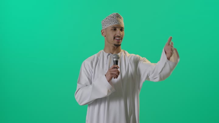 إلقاء المحاضرة وإقامة الندوات، تسجيل المقاطع الصوتية والغنائية، بورتريه لرجل عربي خليجي عماني يرتدي الدشداشة والعمامة يقف يحمل بيده مكبر الصوت، خلفية خضراء