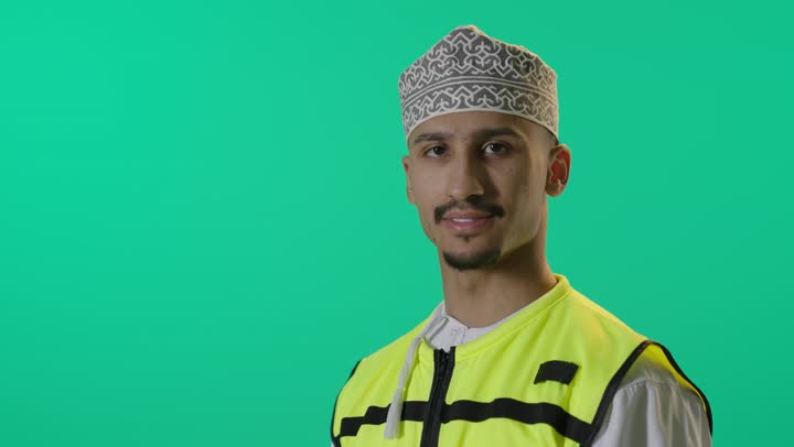 الوقوف بأيدى متقاطعة، بورتريه لمهندس عربي خليجي عماني يرتدي الدشداشة وسترة الحماية ينظر الى الكاميرا فى ثقة، خلفية خضراء
