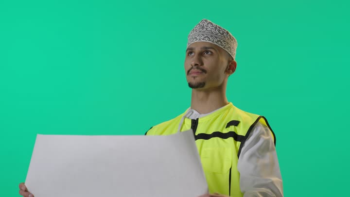 Supervision of architectural engineering projects, auditing project plans, a portrait of an Arab Gulf Omani engineer wearing a dishdasha and a safety vest holding an engineering plan, green background.