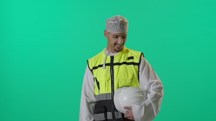 بورتريه لمهندس عربي خليجي عماني يرتدي الدشداشة والكمة وسترة الحماية يحمل بيدة خوذة الحماية ، يقف بشكل مستقيم ينظر إلى الكاميرا بإيماءات الثقة بالنفس، مهنة ووظيفة للشباب في سلطنة عمان، الإشراف على المشاريع الهندسية, مفهوم الانشاءات المعمارية، خلفية خضراء