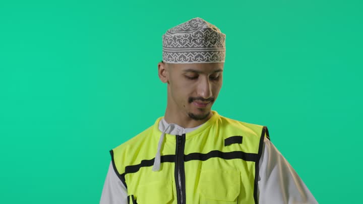 مفهوم الانشاءات المعمارية، بورتريه لمهندس عربي خليجي عماني يرتدي الدشداشة والكمة وسترة الحماية يحمل بيدة خوذة الحماية ، يقف بشكل مستقيم ينظر إلى الكاميرا بإيماءات الثقة بالنفس، مهنة ووظيفة للشباب في سلطنة عمان، الإشراف على المشاريع الهندسية, خلفية خضراء