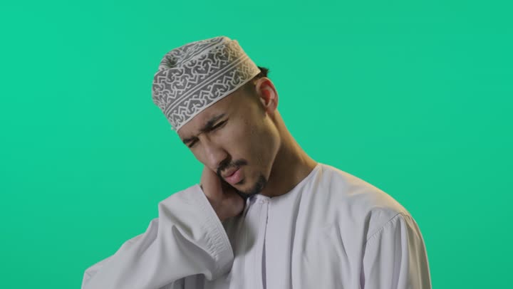 وضع اليد على الرقبة علامة على الوجع الشديد , رجل عربي خليجي عماني يرتدي الدشداشة البيضاء و العمامة , تعابير الشعور بالالم و الاعياء , خلفية خضراء
