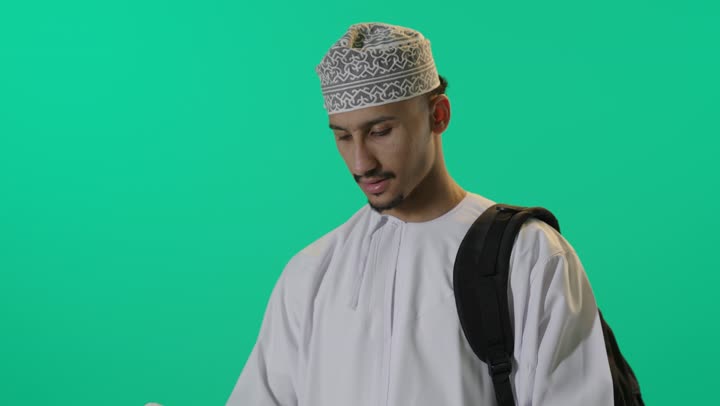 تنمية مهارات القراءة، قضاء أوقات الفراغ بالتثقيف والتعلم, الاهتمام بتصفح الكتب المفيدة, بورتريه لطالب عربي خليجي عماني يرتدي الدشداشة والكمه العمانية حقيبة الظهر يمسك بيده كتاب, خلفية خضراء