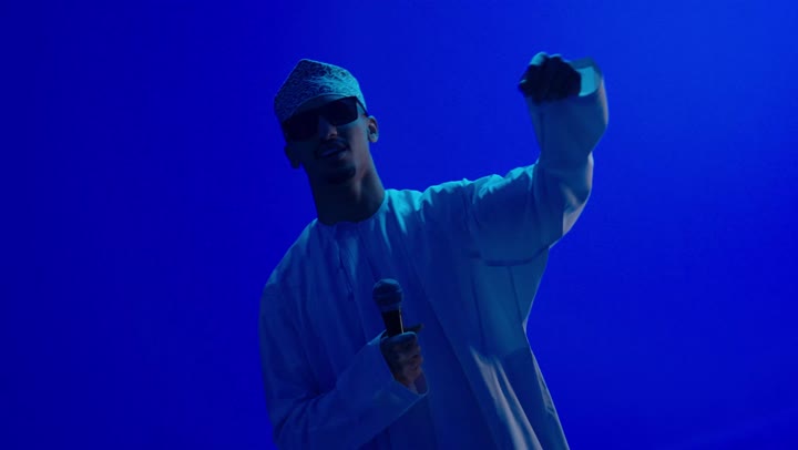 تسجيل مقطع صوتي غنائي, اندماج مع الأغنية، مواهب غنائية فنية, بورتريه لرجل عربي خليجي عماني يرتدي الدشداشة والكمه العمانية و نظارة سوداء يقوم بالغناء باستخدام الميكروفون بتعابير الحماس, خلفية ملونة