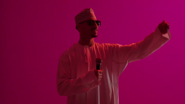 الاندماج مع الأغنية، مواهب غنائية فنية, بورتريه لرجل عربي خليجي عماني يرتدي الدشداشة والكمه العمانية و نظارة سوداء يقوم بالغناء باستخدام الميكروفون بتعابير الحماس, خلفية ملونة
