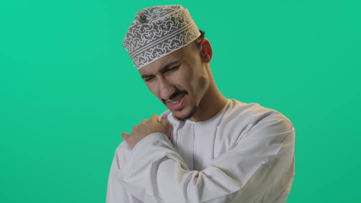 التهاب العضلات والمفاصل, بورتريه لرجل عربي خليجي عماني يرتدي الدشداشة والكمه العمانية يضع يده على كتفه بايماءات الألم والتعب والأرهاق, طلب المساعده والحاجه للعلاج، خلفية خضراء