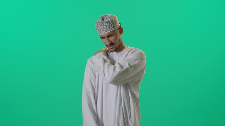 طلب المساعده والحاجه للعلاج، التهاب العضلات والمفاصل, بورتريه لرجل عربي خليجي عماني يرتدي الدشداشة والكمه العمانية يضع يده على كتفه بايماءات الألم والتعب والأرهاق, خلفية خضراء