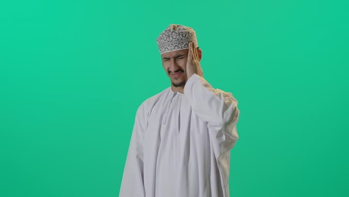 الاحساس بالشقيقية والصداع النصفي, الاجهاد والضغط النفسي,  بورتريه لرجل عربي خليجي عماني يرتدي الدشداشة والكمه العمانية يضع يديه على رأسه ويغمض عينيه بإيماءات الألم الشديد, الإرهاق وفقدان التركيز, الشعور بالصداع وألم الرأس, خلفية خضراء