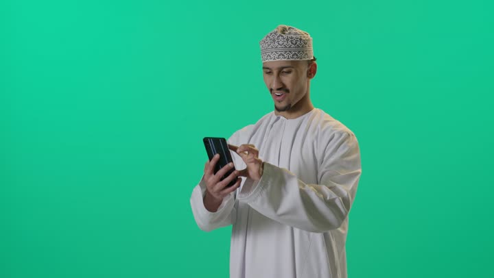 بورتريه لرجل شايب عربي خليجي عماني يرتدي الدشداشة والكمه العمانية يمسك الهاتف المحمول وينظر إليه بإيماءات التركيز والبشاشة، الجسم بالطول الكامل،استخدام الجوال في تصفح مواقع الانترنت، الاستفادة من التقنيات والتكنولوجيا الحديثة، خلفية خضراء