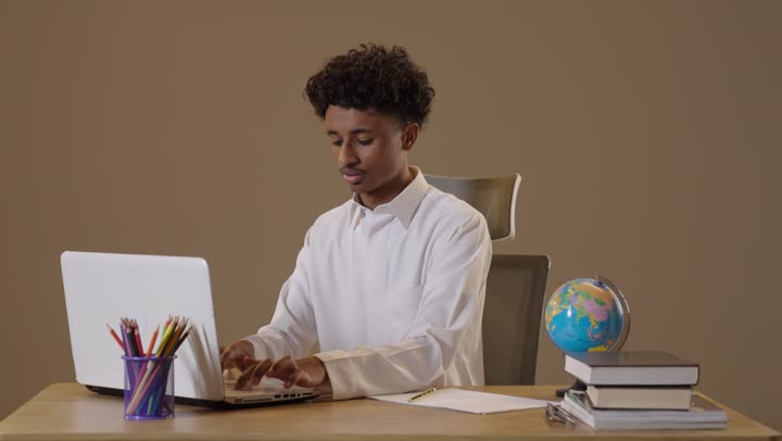A portrait of a Saudi Arabian Gulf Arab student with a dark complexion wearing traditional attire, sitting in his seat and using a computer to solve and review assignments with gestures of joy, utilizing modern technologies in education and remote teaching, back-to-school season, beige background.