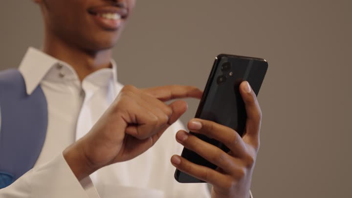 A close-up portrait of a young Arab Gulf Saudi man with a brown complexion, smiling, wearing traditional attire and a backpack, using a mobile phone to browse applications and social media, utilizing modern and advanced devices and technologies, with a beige background.