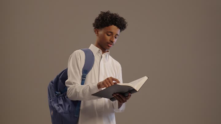 A portrait of a young Arab Gulf Saudi man wearing traditional attire and a backpack reading a book, developing educational curricula in Saudi Arabia, focusing on browsing useful books, preparing for the new school year, reviewing daily lessons, beige background.