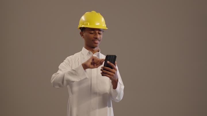 بورتريه لطالب جامعي عربى خليجى سعودى اسمر يرتدي الثوب التقليدى و الخوذة يحمل بيده الهاتف المحمول , دمج التكنولوجيا الحديثة في الأعمال الهندسية, هواية المطالعة والاستكشاف, التواصل مع الأهل والأصدقاء,  خلفية بيج