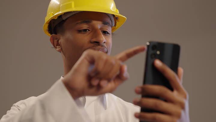 Communication with family and friends, a portrait of an Arab Gulf Saudi engineering university student with a brown complexion wearing traditional attire and a helmet, holding a mobile phone in his hand, integrating modern technology in engineering work, a hobby of reading and exploration with a beige background.