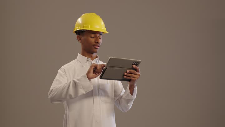 بورتريه لطالب جامعي عربى خليجى سعودى اسمر  يرتدي الثوب التقليدى و الخوذة يحمل بيده جهاز التابلت المحمول , دمج التكنولوجيا الحديثة في الأعمال الهندسية, هواية المطالعة والاستكشاف خلفية بيج