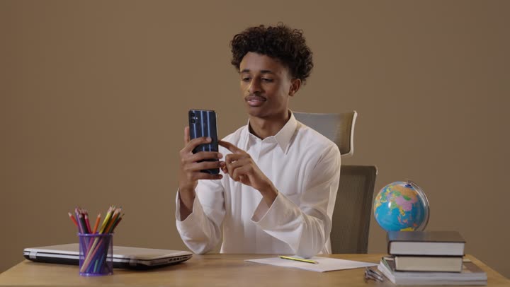 Integrating technology with education, the concept of distance learning, attending online classes via the internet, a portrait of a dark-skinned Arab Gulf Saudi student wearing traditional attire sitting in his seat and using his phone to solve and review assignments with gestures of joy, beige background.