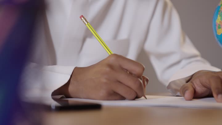 مجسم الكرة الأرضية على طاولة مكتبية، التعليم والتدريس في مدارس السعودية، موسم العودة للمدرسة، بورتريه لطالب عربي خليجي سعودي يرتدي الثوب التقليدي يجلس في مقعده ويقوم بمراجعة الدروس وحل الواجبات, خلفية بيج