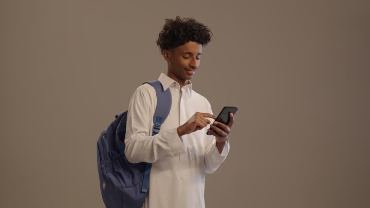 البحث والمطالعة من خلال الجوال، تصفح التطبيقات ووسائل التواصل الاجتماعي، استخدام الأجهزة والتقنيات الحديثة والمتطورة, بورتريه لشاب عربي خليجي سعودي اسمر مبتسم يرتدي الثوب التقليدي  و حقيبة ظهر يستخدم الهاتف المحمول, خلفية بيج