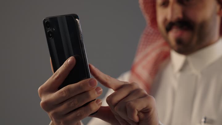 بورتريه مقرب لرجل عربي خليجي سعودي يرتدي الثوب التقليدى و الشماغ يستخدم الجوال, تصفح الانترنت والتسوق الالكتروني، استخدام الهاتف المحمول في التواصل مع الأفراد، دمج العمل مع التقنية والتكنولوجيا, خلفية رمادية