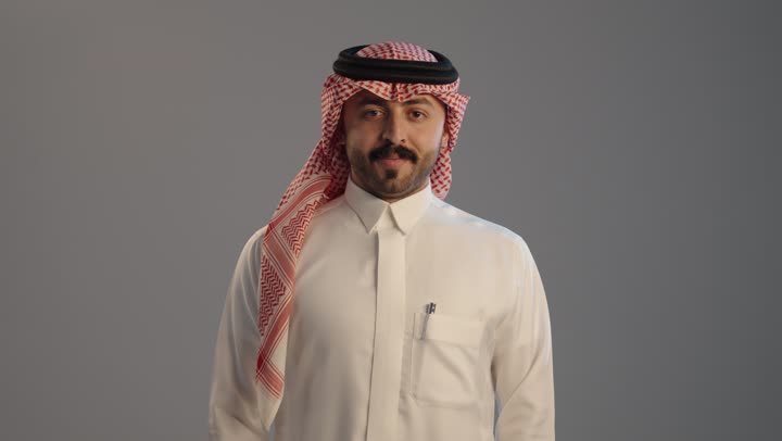 بورتريه مقرب لرجل عربي خليجي سعودي يرتدي الثوب التقليدي والشماغ ويرفع أصابع الإبهام لأعلى، إيماءات السعادة والإعجاب والموافقة، الإمتياز والتميز والنجاح, حركة أصبع تدل على القبول والرضا, خلفية رمادية