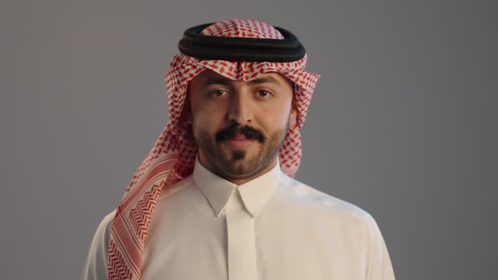 الإمتياز والتميز والنجاح، بورتريه مقرب لرجل عربي خليجي سعودي يرتدي الثوب التقليدي والشماغ ويرفع أصابع الإبهام لأعلى، إيماءات السعادة والإعجاب والموافقة، حركة أصبع تدل على القبول والرضا, خلفية رمادية