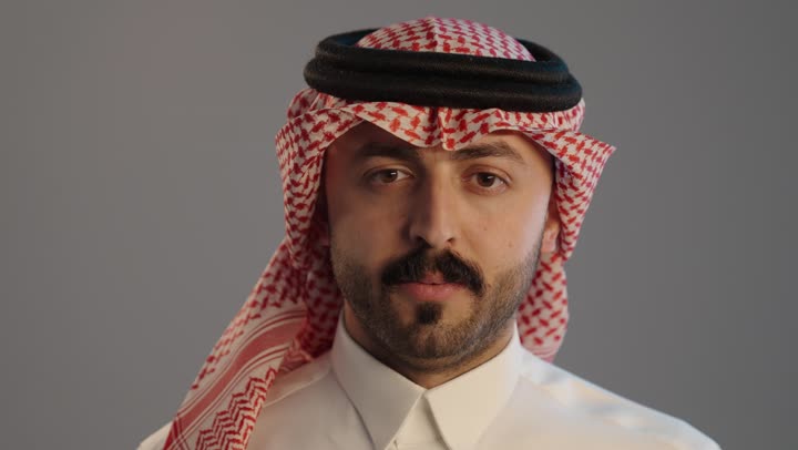 بورتريه لمواطن عربي خليجي سعودي يرتدي الثوب التقليدي والشماغ ويضع أصبع السبابة على فمه، الهدوء والسكون وعدم الإزعاج، التوقف عن الكلام وخفض الصوت، إيماءات وجه ويدين تدل على طلب السكوت والصمت، حركة أصبع وإشارات, خلفية رمادية
