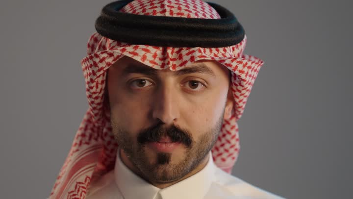 التوقف عن الكلام وخفض الصوت، بورتريه لمواطن عربي خليجي سعودي يرتدي الثوب التقليدي والشماغ ويضع أصبع السبابة على فمه، الهدوء والسكون وعدم الإزعاج، إيماءات وجه ويدين تدل على طلب السكوت والصمت، حركة أصبع وإشارات, خلفية رمادية