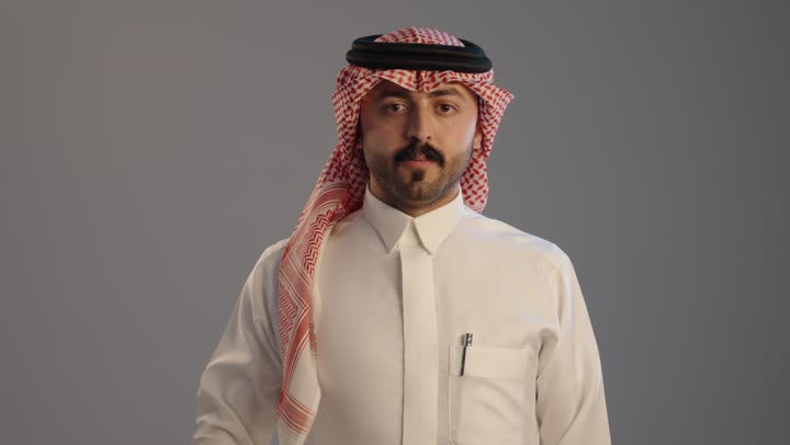 A portrait of a Saudi Arabian Gulf man wearing traditional attire and a shemagh, raising his hand upwards in a stop gesture, showing signs of fear, anxiety, and discomfort about something, expressing disapproval and rejection of harm, requesting to stop and refrain from doing something, with a gray background.