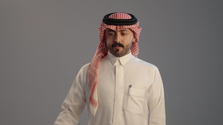 بناء جسم رياضي، مفهوم اللياقة البدنية و كمال الأجسام، بورتريه مقرب لرجل عربي خليجي سعودي يرتدي الثوب التقليدى و الشماغ  يقوم بعرض عضلاته, الشعور بالقوة والسرور, خلفية رمادية