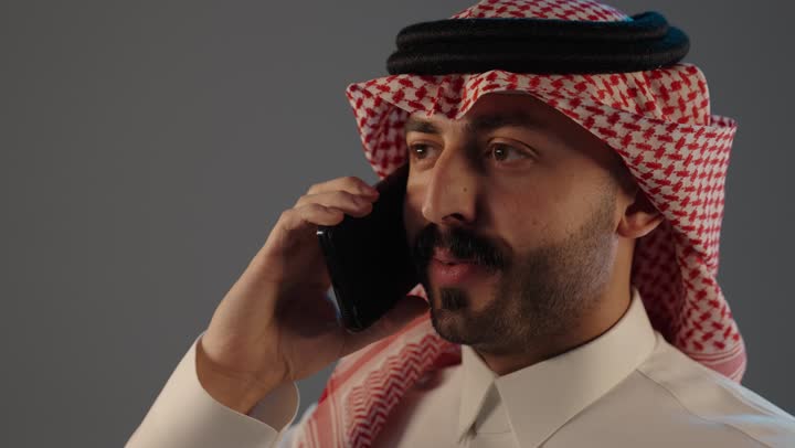 استخدام جهاز تقنى حديث, تبادل الحديث مع الأهل والأصدقاء, بورتريه مقرب لرجل عربي خليجي سعودي يرتدي الثوب التقليدى و الشماغ يستخدم الهاتف, اجراء مكالمة هاتفية صوتية عبر الجوال, مفهوم ادارة الاعمال عن بعد, خلفية رمادية