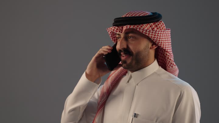 تبادل الحديث مع الأهل والأصدقاء, بورتريه مقرب لرجل عربي خليجي سعودي يرتدي الثوب التقليدى و الشماغ يستخدم الهاتف, اجراء مكالمة هاتفية صوتية عبر الجوال, مفهوم ادارة الاعمال عن بعد, استخدام جهاز تقنى حديث, خلفية رمادية