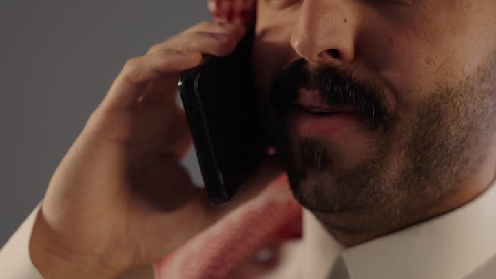 بورتريه مقرب لرجل عربي خليجي سعودي يرتدي الثوب التقليدى و الشماغ يستخدم الهاتف, إيماءات السعادة بالتواصل مع الأصدقاء والأقارب, إجراء مكالمة هاتفية صوتية عبر الجوال, خلفية رمادية