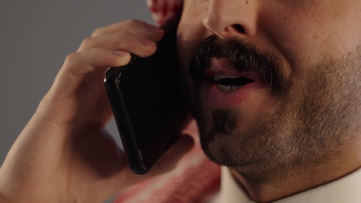 إجراء مكالمة هاتفية صوتية عبر الجوال, بورتريه مقرب لرجل عربي خليجي سعودي يرتدي الثوب التقليدى و الشماغ يستخدم الهاتف, إيماءات السعادة بالتواصل مع الأصدقاء والأقارب, خلفية رمادية