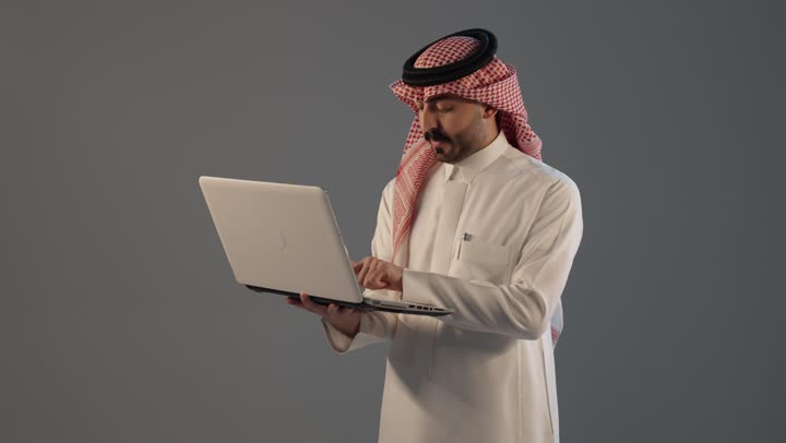 دمج العمل مع التقنية والتكنولوجيا, بورتريه لرجل عربي خليجي سعودي يرتدي الثوب التقليدى و الشماغ يعمل على الحاسوب المحمول, النقر على لوحة المفاتيح، إدارة شؤون الأعمال وتنظيمها عبر الأجهزة التقنية، خلفية رمادية