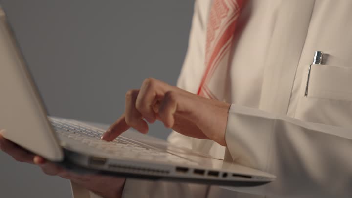 بورتريه مقرب ليد رجل عربي خليجي سعودي يرتدي الثوب التقليدى يعمل على الحاسوب المحمول, دمج العمل مع التقنية والتكنولوجيا, النقر على لوحة المفاتيح، إدارة شؤون الأعمال وتنظيمها عبر الأجهزة التقنية، خلفية رمادية