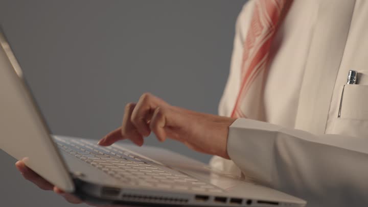 النقر على لوحة المفاتيح، إدارة شؤون الأعمال وتنظيمها عبر الأجهزة التقنية، بورتريه مقرب ليد رجل عربي خليجي سعودي يرتدي الثوب التقليدى يعمل على الحاسوب المحمول, دمج العمل مع التقنية والتكنولوجيا, خلفية رمادية