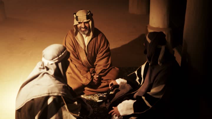 An elderly Arab Saudi Gulf man wearing a bisht and agal on Foundation Day sits with two men inside a mud building, commemorating the establishment of the first Saudi state by Imam Muhammad bin Saud, on the day we began, February 22, 1727. The historical and cultural depth of the Kingdom, wearing traditional attire reflects the heritage.