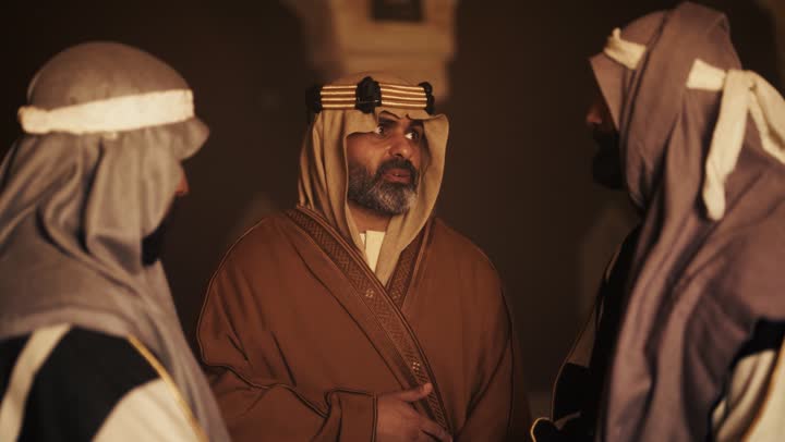 رجل كبير السن عربي سعودي خليجي يرتدي البشت وعقال يوم التأسيس يجلس مع رجلين داخل مبنى طيني, ذكرى تأسيس الدوله السعوديه الاولى على يد الامام محمد بن سعود, يوم بدينا 22 فبراير 1727م, العمق التاريخي والحضاري للمملكة، ارتداء الأزياء التقليدية تعكس التراث