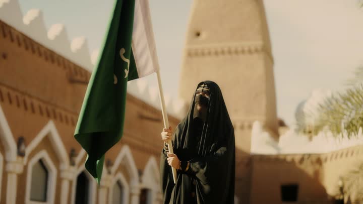 Wearing traditional folk costumes, a symbol of national identity and the banner of Islam and monotheism, on the day we began in 1727 AD, the old heritage clay houses, an Arab Gulf Saudi woman wearing a black abaya and a niqab holding the founding flag, the anniversary of the establishment of the first Saudi state on February 22.