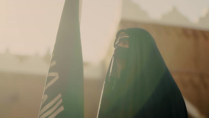 The logo of national identity and the banner of Islam and monotheism, the day we began in 1727 AD, wearing traditional folk costumes, the old heritage clay houses, a close-up shot of a Saudi Gulf Arab woman wearing a black abaya and niqab holding the founding flag, the anniversary of the establishment of the first Saudi state on February 22.