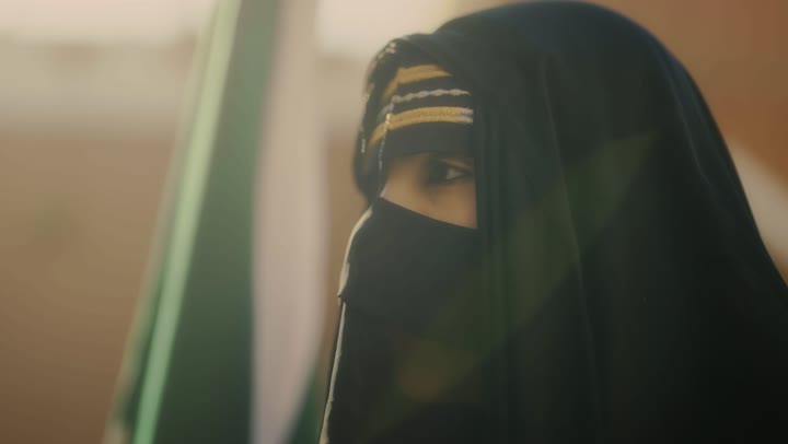 يوم بدينا 1727 م، ارتداء الأزياء الشعبية التقليدية، البيوت الطينية التراثية القديمة، شعار الهوية الوطنية وراية الإسلام والتوحيد، تصوير مقرب لامرأة عربية خليجية سعودية ترتدي العباءة السوداء والبرقع تحمل علم التأسيس, ذكرى تأسيس الدولة السعودية الأولى 22 فبراير