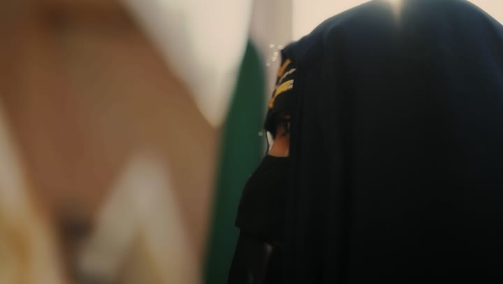 The logo of national identity and the banner of Islam and monotheism, the day we began in 1727 AD, wearing traditional folk costumes, the old heritage clay houses, a close-up shot of a Saudi Gulf Arab woman wearing a black abaya and niqab holding the founding flag, the anniversary of the establishment of the first Saudi state on February 22.