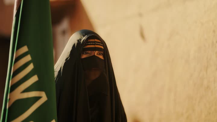 On the day we began in 1727 AD, wearing traditional folk costumes, the old heritage mud houses, the emblem of national identity and the flag of Islam and monotheism, a close-up shot of a Saudi Gulf Arab woman wearing a black abaya and a niqab holding the founding flag, commemorating the establishment of the first Saudi state on February 22.