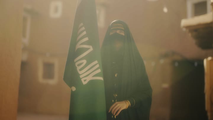 ارتداء الأزياء الشعبية التقليدية، شعار الهوية الوطنية وراية الإسلام والتوحيد، يوم بدينا 1727 م، البيوت الطينية التراثية القديمة، امرأة عربية خليجية سعودية ترتدي العباءة السوداء والبرقع تحمل علم التأسيس, ذكرى تأسيس الدولة السعودية الأولى 22 فبراير