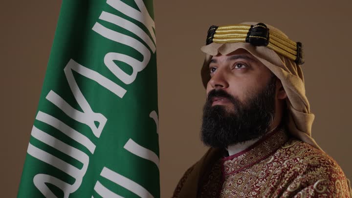 رجل سعودي يرتدي لباس يوم التأسيس بجانب العلم السعودي