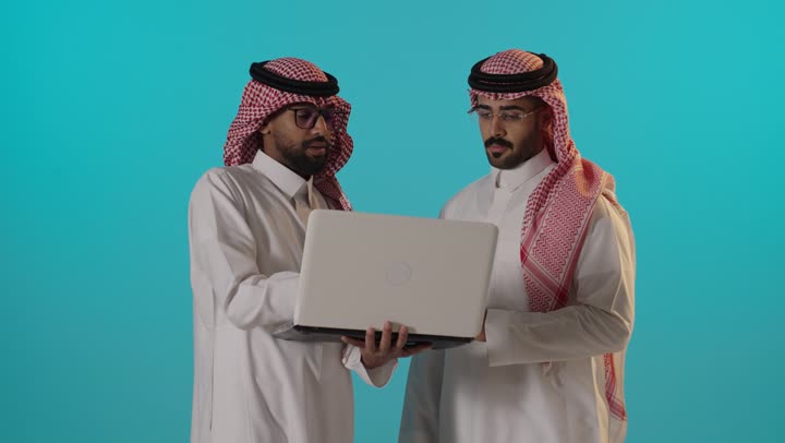 رجلان سعوديان يناقشان العمل باستخدام كمبيوتر محمول بالاستوديو