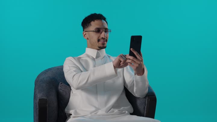 رجل سعودي بالثوب يستخدم الهاتف الذكي بخلفية زرقاء